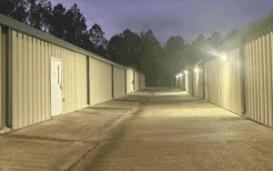 Well-lit concrete drive aisle between beige self-storage buildings at night featuring security lighting and a gated perimeter fence.