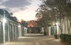 Paved drive aisle at a self-storage facility at dusk, lined with exterior security lights and drive-up units with green roll-up doors.